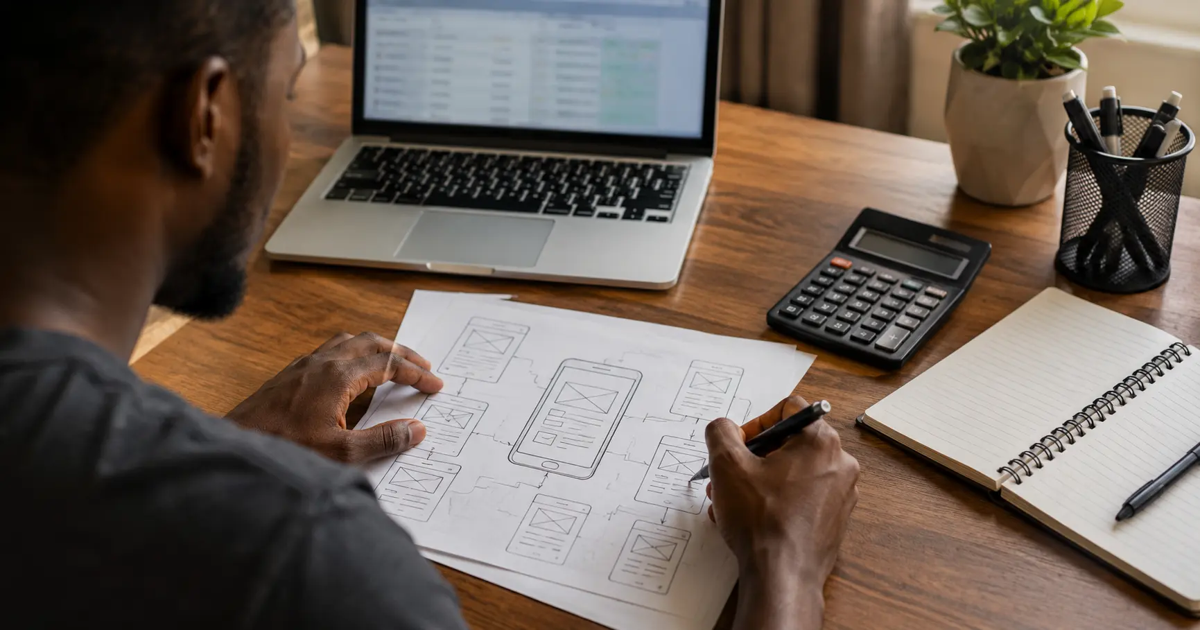Developer sketching an app wireframe with a calculator and laptop on desk, planning app development budget